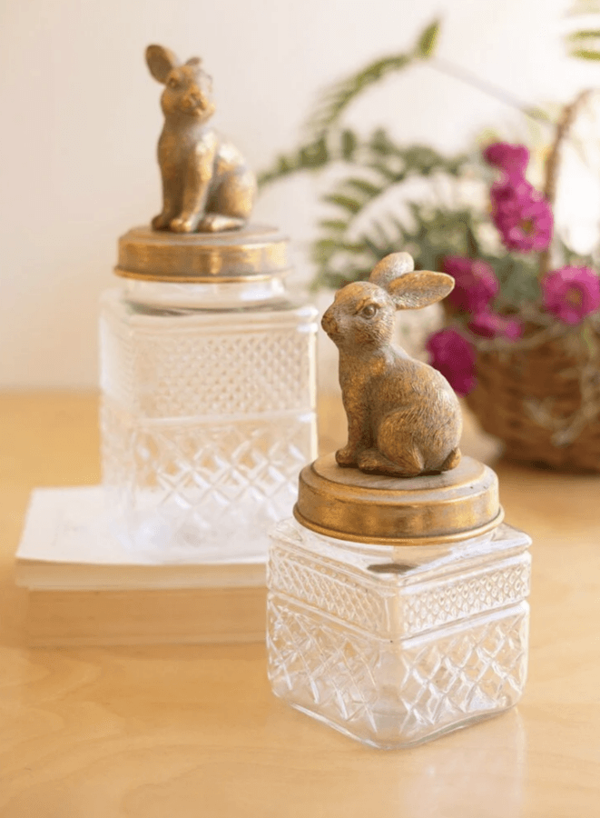 19 Fresh FINDS Around BHAM: Closet Refreshers, Entertaining Picks & More - 13 Two clear glass jars with ornate lids featuring gold rabbit figurines, placed on a wooden surface in Birmingham—perfect fresh finds with a book and a basket of flowers in the background.