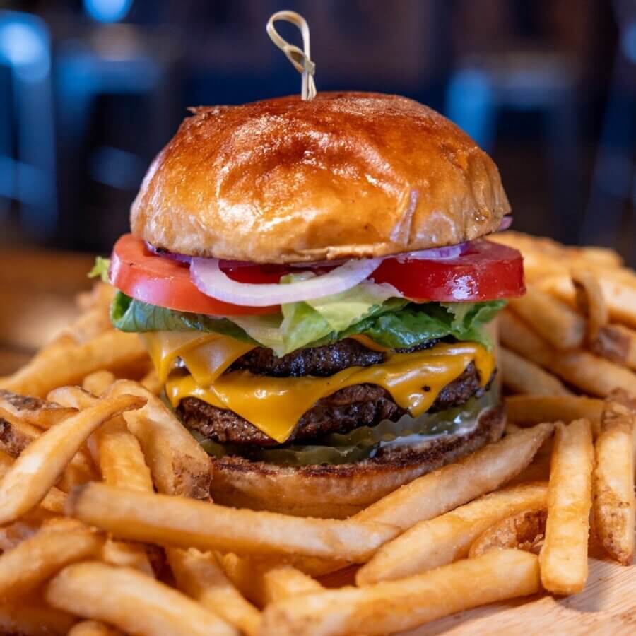 11 Must-Try Burgers in Birmingham - 3 A must-try cheeseburger in BHAM, stacked with lettuce, tomato, and onion on a brioche bun, served alongside crispy French fries on a wooden surface at Back Forty.