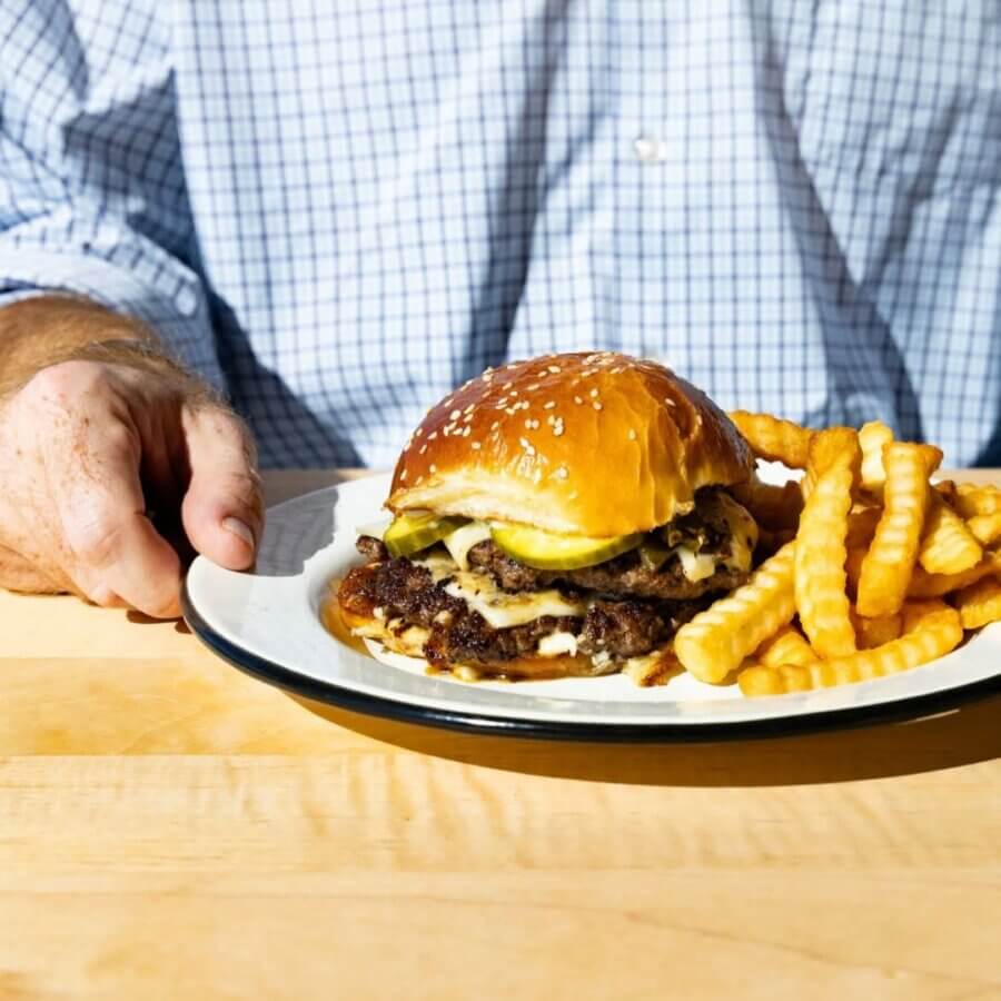 11 Must-Try Burgers in Birmingham - 6 A person in a checkered shirt sits at a table in BHAM with a plate holding a must-try cheeseburger topped with pickles and a serving of crinkle-cut fries at Hero Diner.