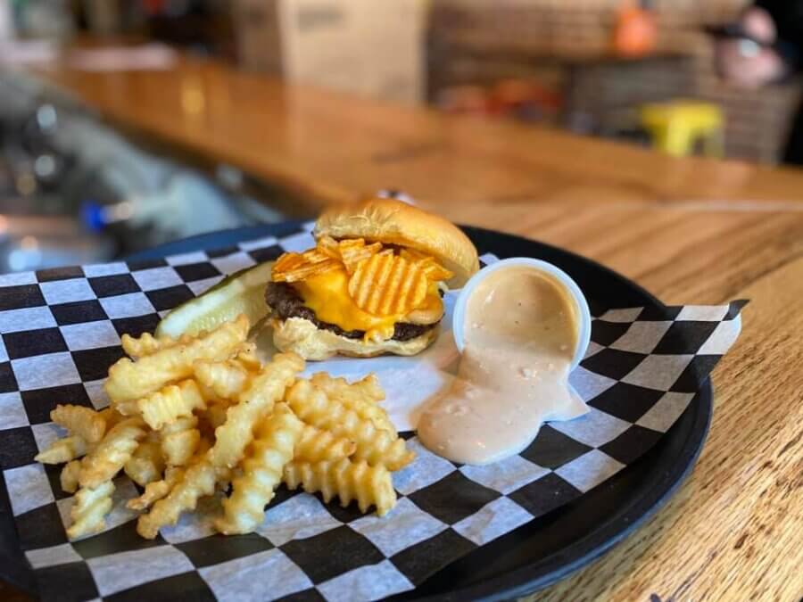 11 Must-Try Burgers in Birmingham - 4 A must-try cheeseburger in BHAM, topped with potato chips, crinkle-cut fries, a pickle spear, and a side of dipping sauce—all served on a checkered paper-lined black plate at Jack Brown's Beer and Burger Joint.