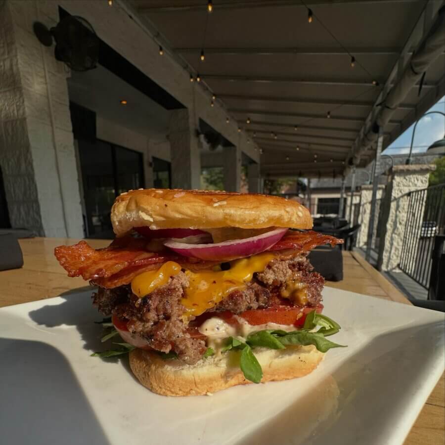 11 Must-Try Burgers in Birmingham - 11 A close-up of a must-try cheeseburger with bacon, lettuce, tomato, red onion, and sauce on a white plate, set on an outdoor restaurant table at Porch.