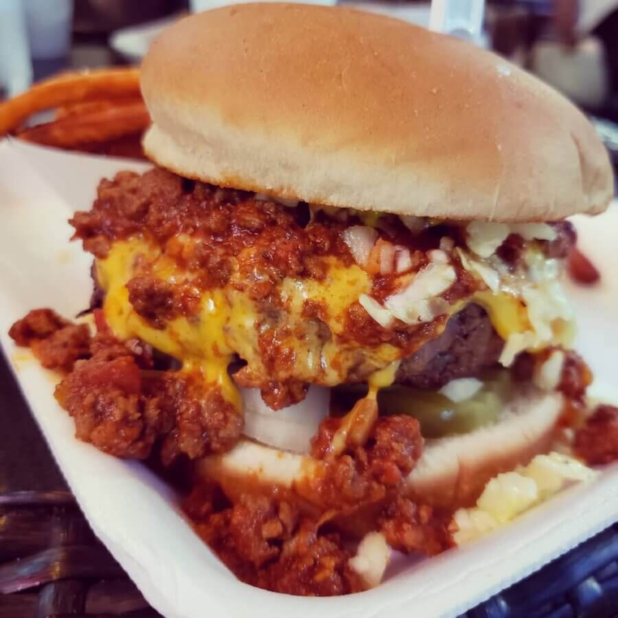 11 Must-Try Burgers in Birmingham - 8 A close-up of a BHAM cheeseburger topped with chili, onions, and melted cheese in a takeout container at Saw's Juke Joint.