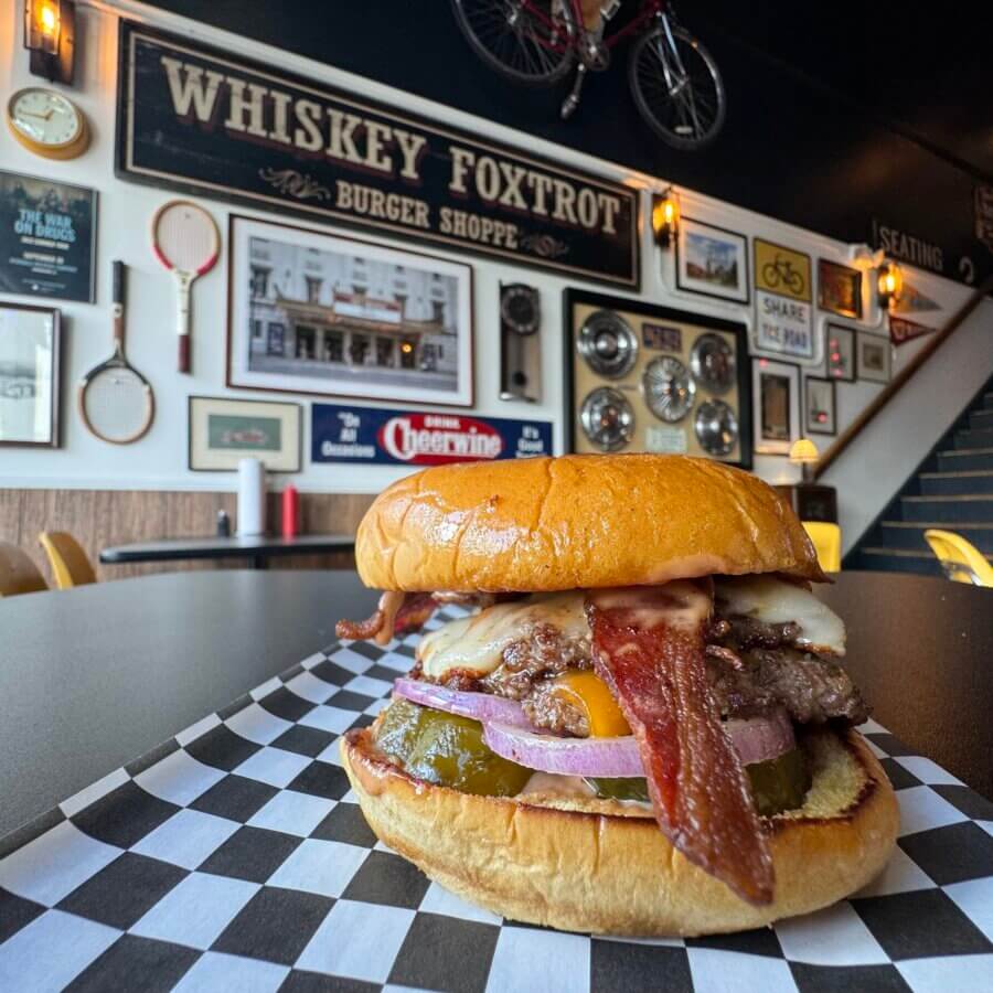 11 Must-Try Burgers in Birmingham - 10 Close-up of a must-try bacon cheeseburger with onions and pickles at Whiskey Foxtrot Burger Shoppe, one of BHAM’s best spots for burgers, served on checkered paper with framed decor visible in the background.