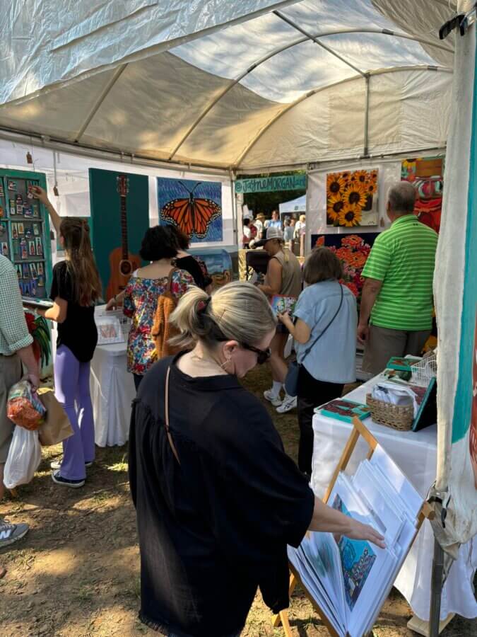 Your Guide to BHAM's Bluff Park - 9 People browse artwork and prints at the Bluff Park outdoor art market under a white tent. Tables display colorful paintings and crafts, while a guide offers insight into each piece. The ground is covered with dirt and scattered leaves.