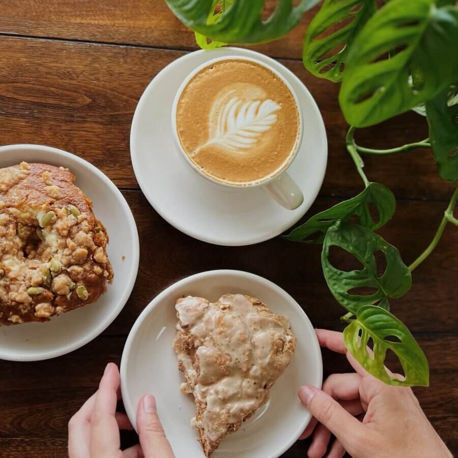 Your Guide to BHAM's Bluff Park - 4 At Bluff Park Coffee Collective, a cup of latte with latte art, a scone, and a muffin sit on a wooden table next to a green plant. Two hands hold the plate with the scone.