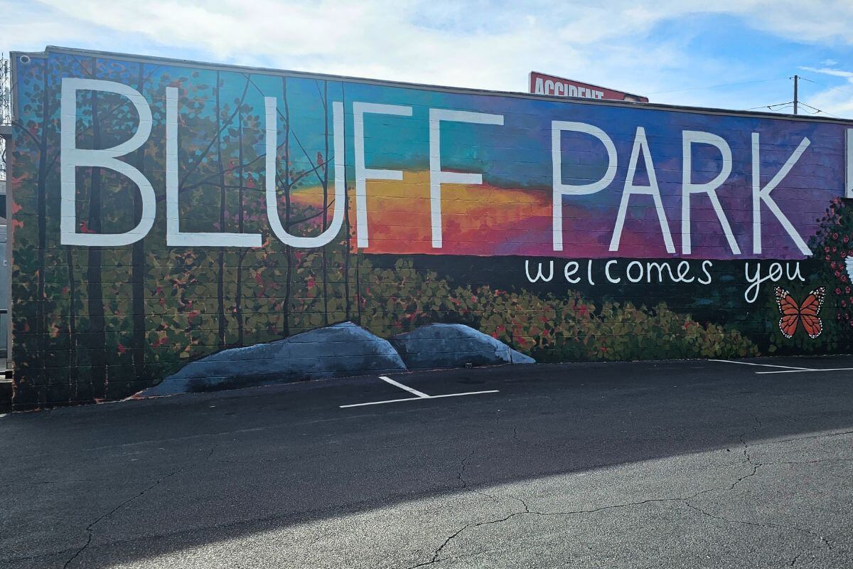 A mural in Bluff Park reads "BLUFF PARK welcomes you," featuring a landscape, trees, sunset, and butterfly; an empty parking lot lies in front—a charming scene for any Bluff Park travel guide.