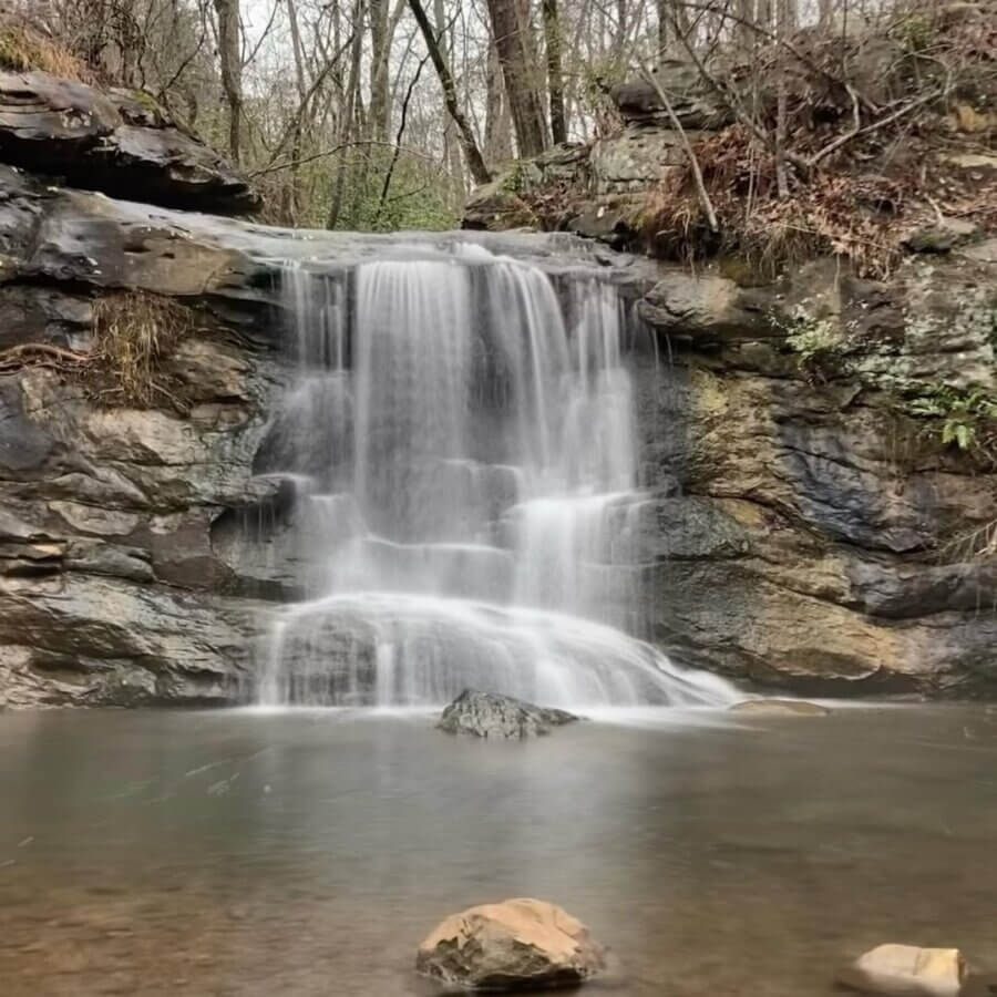 Your Guide to BHAM's Bluff Park - 12 A small waterfall flows over rocky ledges into a calm pool, surrounded by trees and foliage—an inviting scene in Bluff Park that nature lovers will cherish.