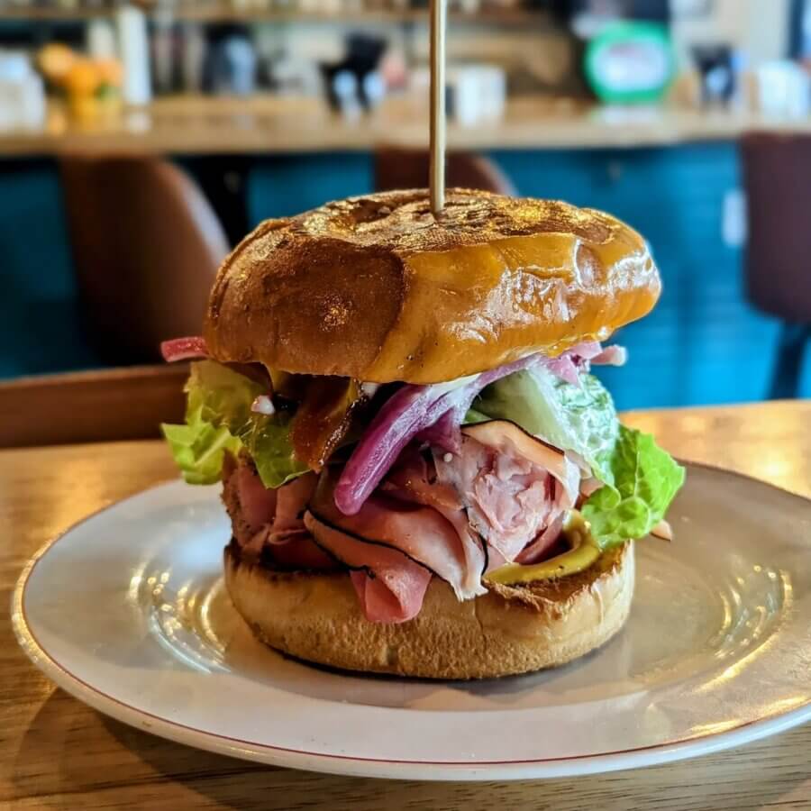Your Guide to BHAM's Bluff Park - 6 A sandwich with ham, lettuce, pickles, and onions served on a toasted bun, secured with a skewer, sits on a white plate—a tasty find for any Bluff Park travel adventure in the heart of the restaurant scene.