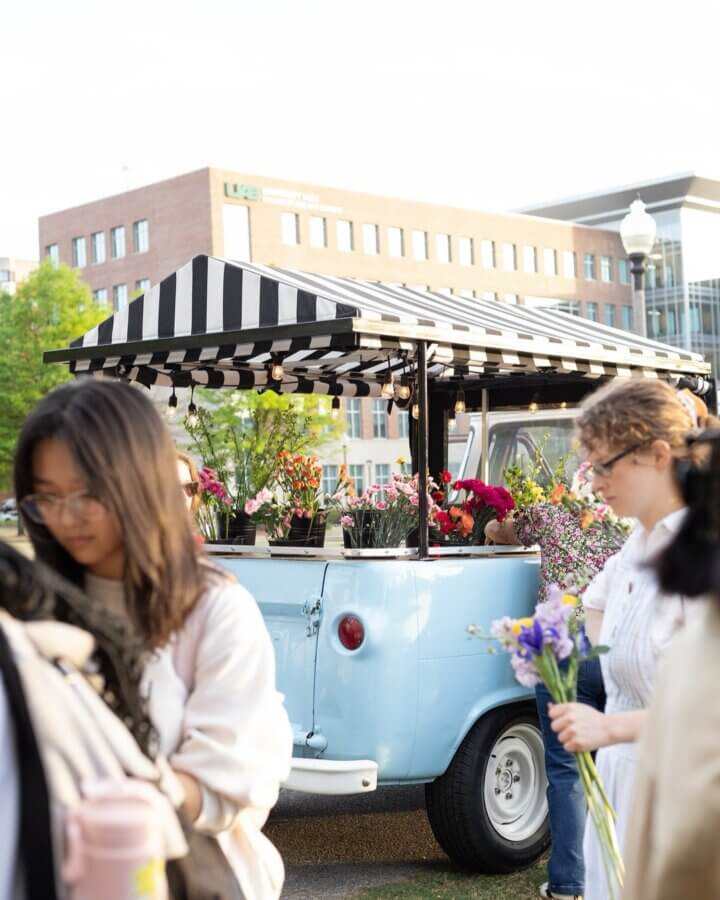 Your Guide to BHAM's Bluff Park - 13 People gather around a light blue flower truck with a black-and-white striped canopy in Bluff Park, selecting bouquets as the sunshine guides their day, with buildings in the background.