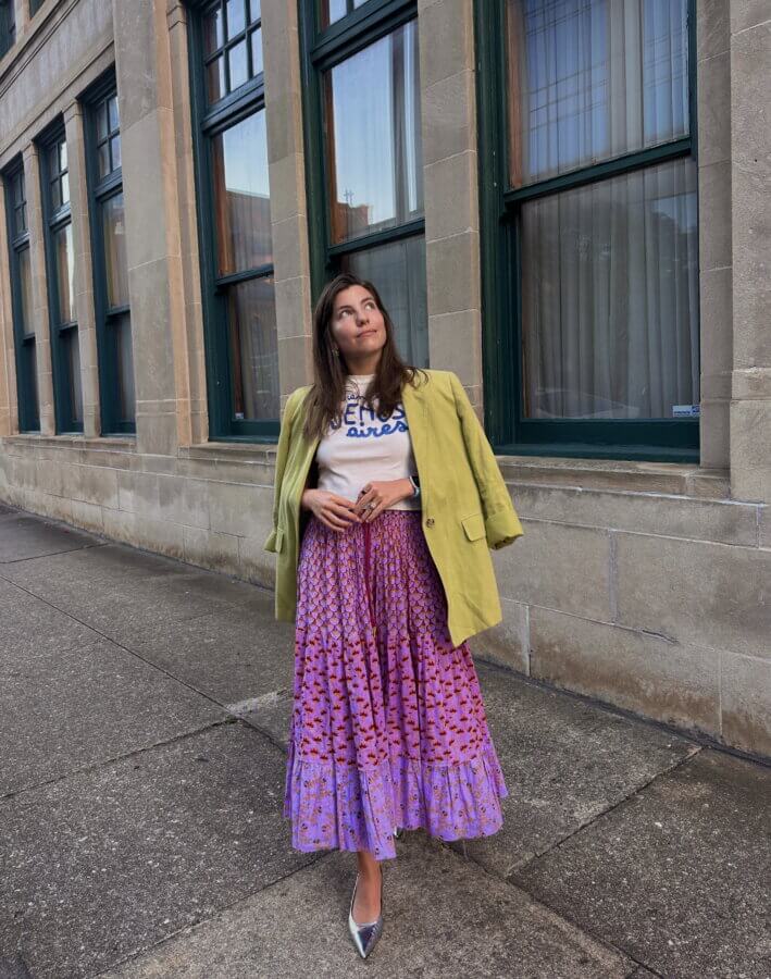 She’s Behind “One Week in Birmingham” - 2 Hattie O'Hara stands on a sidewalk near a building, dressed in a green blazer, graphic t-shirt, purple patterned skirt, and silver shoes. She looks up thoughtfully, as if reflecting on her adventures during One Week in Birmingham.