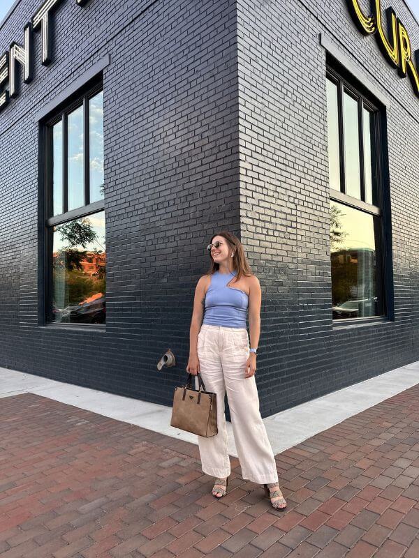 She’s Behind “One Week in Birmingham” - 3 A woman in a blue tank top, beige pants, and sandals stands on a brick sidewalk with her tan handbag outside a black brick building with large windows.