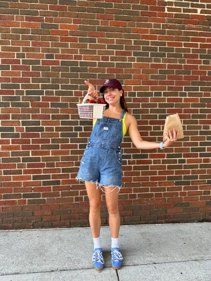 She’s Behind “One Week in Birmingham” - 4 Hattie O'Hara in denim overalls and a maroon cap stands against a brick wall, holding a basket of fruit and a brown paper bag.