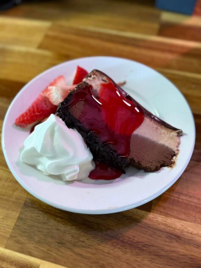 10 New Birmingham Restaurants to Try This Month - 4 A slice of chocolate and vanilla cheesecake topped with cherry sauce, served with whipped cream and sliced strawberries on a white plate.