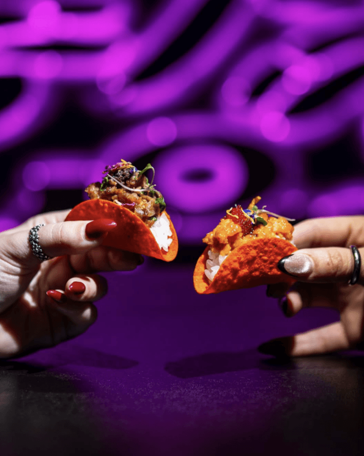 10 New Birmingham Restaurants to Try This Month - 9 Two hands holding mini tacos with orange shells and assorted fillings against a blurred background with purple neon lights—a delicious find among new restaurants to try in Birmingham.