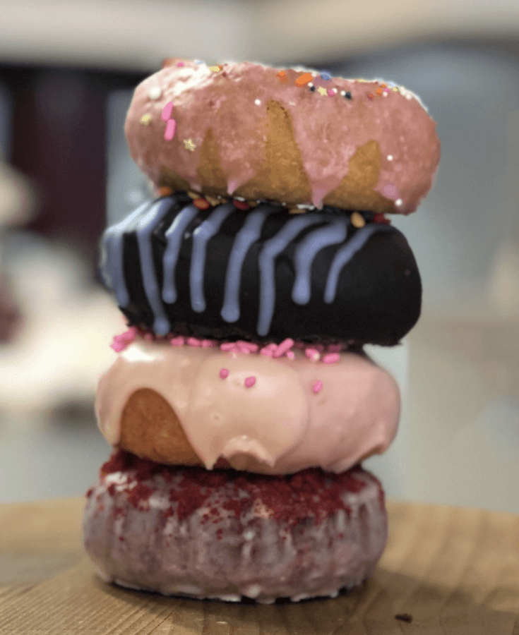 10 New Birmingham Restaurants to Try This Month - 10 A stack of four assorted frosted donuts, each with different icing and sprinkles, placed on a wooden surface—a sweet highlight among new restaurants in the Birmingham dining scene.