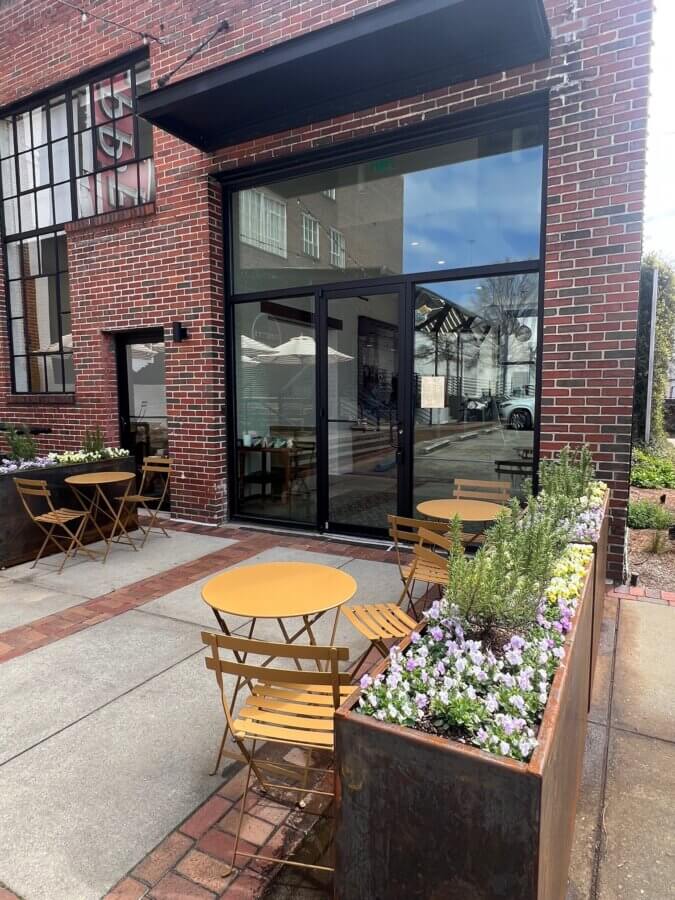 10 New Birmingham Restaurants to Try This Month - 7 Outdoor seating area with yellow tables and chairs on a sidewalk, next to flower planters, outside a brick building with large glass doors—perfect for enjoying one of the newest Birmingham restaurants or discovering unique places to eat.
