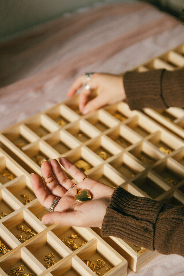 She’s Behind a BHAM Charm Bar, Craft Club, AND a Book Bus! - 5 A person with rings on their fingers selects a gold pendant from the Charm Bar—a wooden display tray filled with various gold jewelry pieces at Craft Club BHAM.