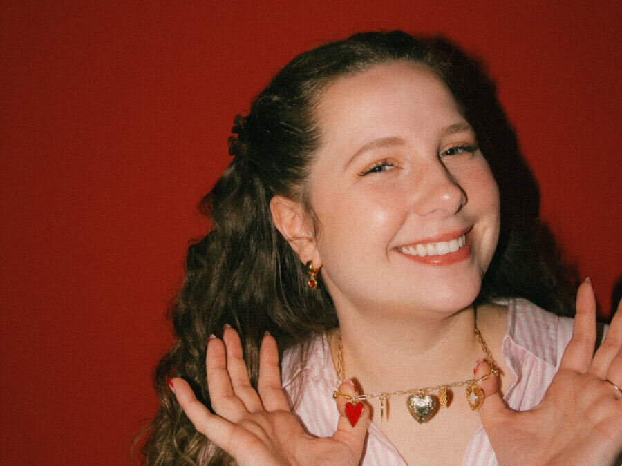 She’s Behind a BHAM Charm Bar, Craft Club, AND a Book Bus! - 2 Rachael Welch smiles and holds up a gold necklace with heart charms from the BHAM Charm Bar in front of a red background.