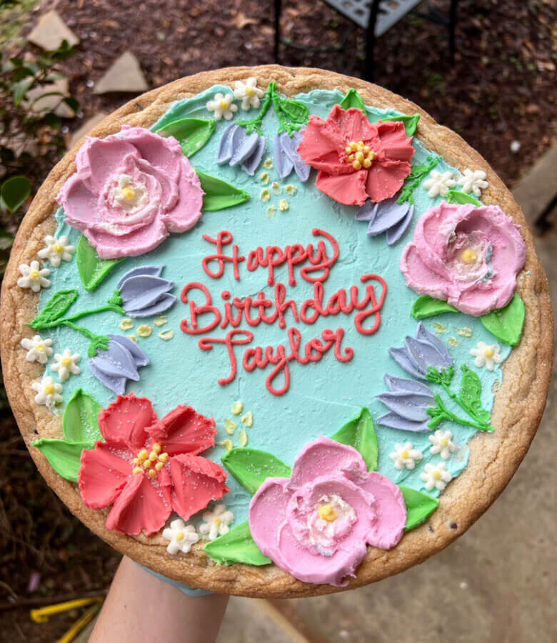 The Baker Everyone in Bluff Park Is Talking About - 3 A cookie cake from Good Neighbor Baking, decorated with pastel flowers and leaves, features the message "Happy Birthday Taylor" written in red icing on a light blue background.