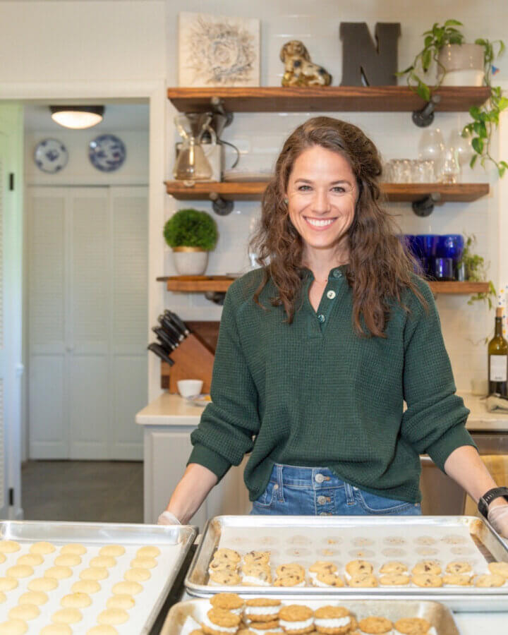 The Baker Everyone in Bluff Park Is Talking About - 2 Ramsey Nuss stands in a kitchen smiling at the camera with several trays of cookies in front of her on the counter, proudly representing Good Neighbor Baking.
