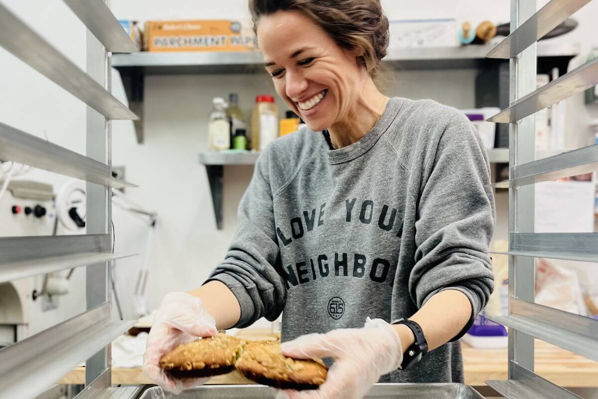 Ramsey Nuss wearing gloves and a "Love Your Neighbor" sweatshirt smiles while placing cookies on a baking tray in a kitchen, embodying the spirit of Good Neighbor Baking.