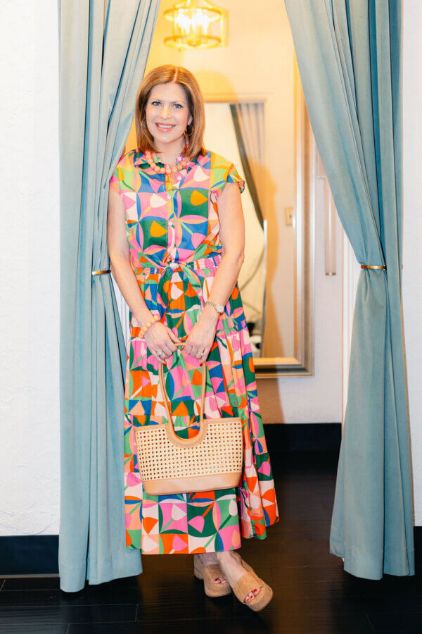 3 BHAM Boutiques Share Their Favorite Spring 2026 Looks - 4 A woman stands between blue curtains, wearing a colorful geometric-patterned dress from BHAM Boutiques, holding a woven handbag, and smiling at the camera—one of our favorite spring looks.