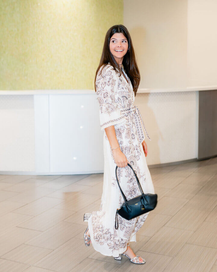 3 BHAM Boutiques Share Their Favorite Spring 2026 Looks - 10 A woman in a long, patterned white dress and sandals stands indoors, holding a black handbag and smiling while looking to her left—one of our favorite spring outfits from BHAM Boutiques.