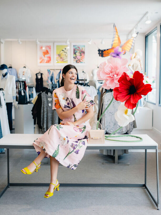 3 BHAM Boutiques Share Their Favorite Spring 2026 Looks - 7 A woman in a floral dress sits on a table in a clothing store, smiling, with oversized artificial flowers and a butterfly decoration beside her.