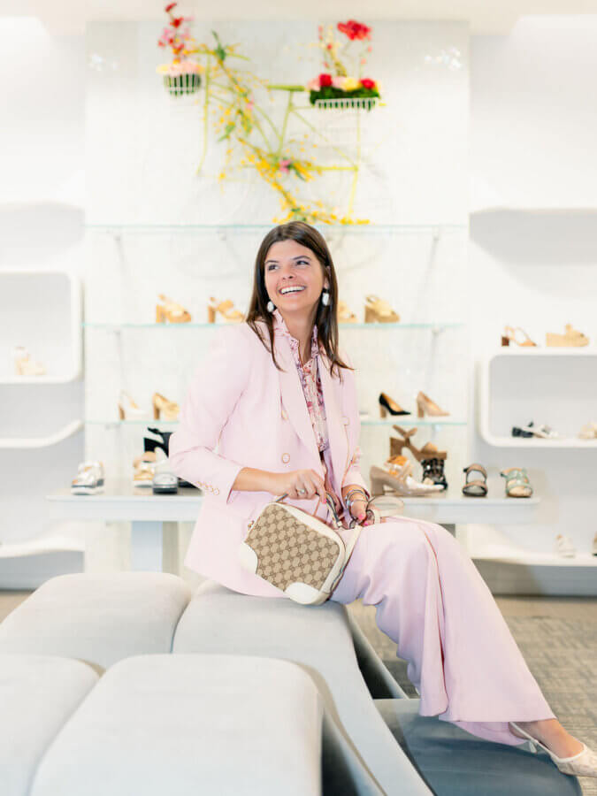 3 BHAM Boutiques Share Their Favorite Spring 2026 Looks - 9 A woman in a light pink suit sits on a cushioned bench, holding a designer handbag—one of our favorite Spring Looks from BHAM Boutiques—surrounded by shelves of shoes and a bicycle wall decoration behind her.