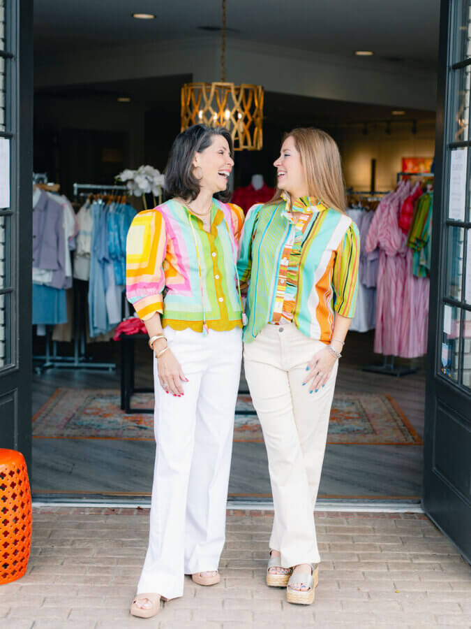 3 BHAM Boutiques Share Their Favorite Spring 2026 Looks - 14 Two women stand smiling at each other in the entrance of a clothing store, both showcasing vibrant boutique fashion with colorful patterned tops and light-colored pants. Clothing racks are visible in the background.