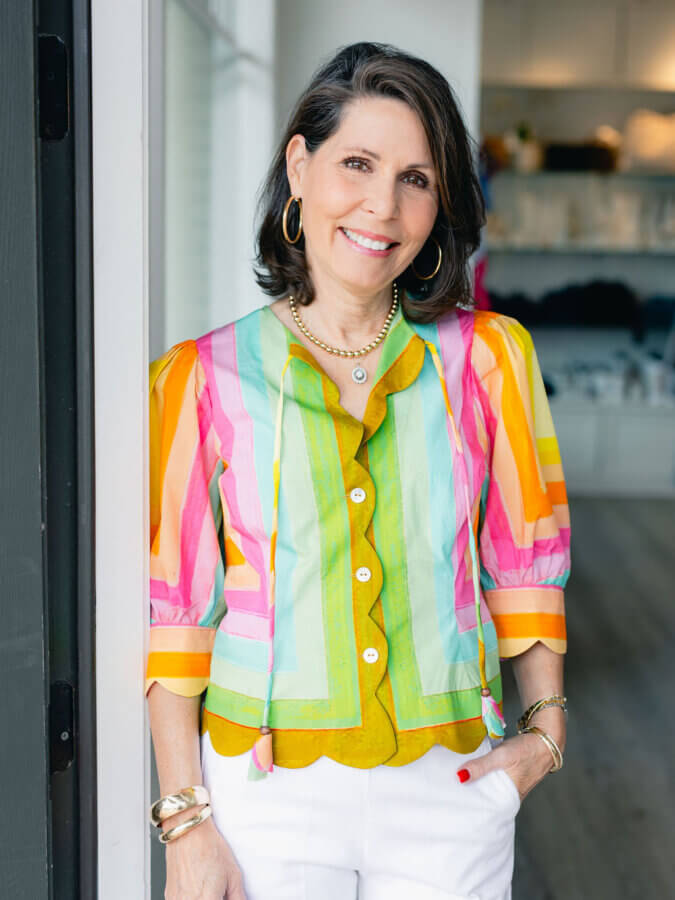 3 BHAM Boutiques Share Their Favorite Spring 2026 Looks - 16 Woman with shoulder-length dark hair wearing a bright, multicolored blouse and white pants, standing indoors and smiling at the camera—one of our Favorite Spring Looks from BHAM Boutiques.