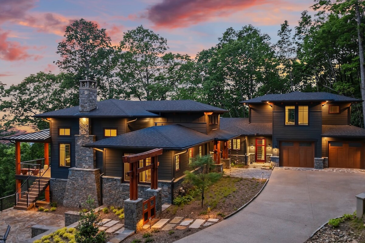 Modern two-story house with stone and wood accents, large windows, and exterior lights on, set against trees and a colorful sunset sky—an inspiring showcase of custom home design services.