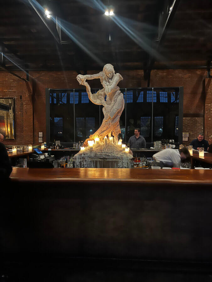 Your Guide to Chattanooga's Culinary Scene - 2 A large sculpture of a woman holding an object is displayed behind a bar with candles in a dimly lit Chattanooga venue; bartenders work in the background.