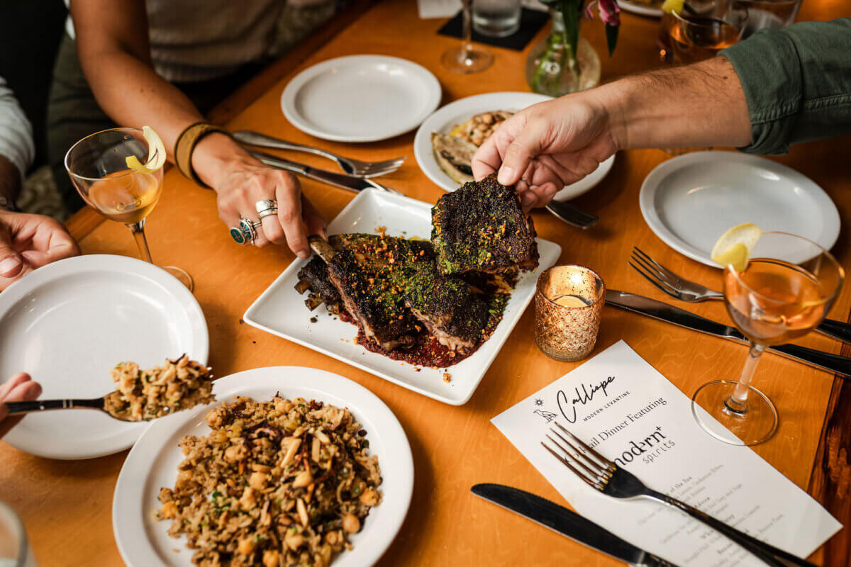 A group shares a meal at a restaurant, with hands reaching for grilled meat on a platter, drinks, plates, utensils, and a menu on the table—true dining in Chattanooga style.