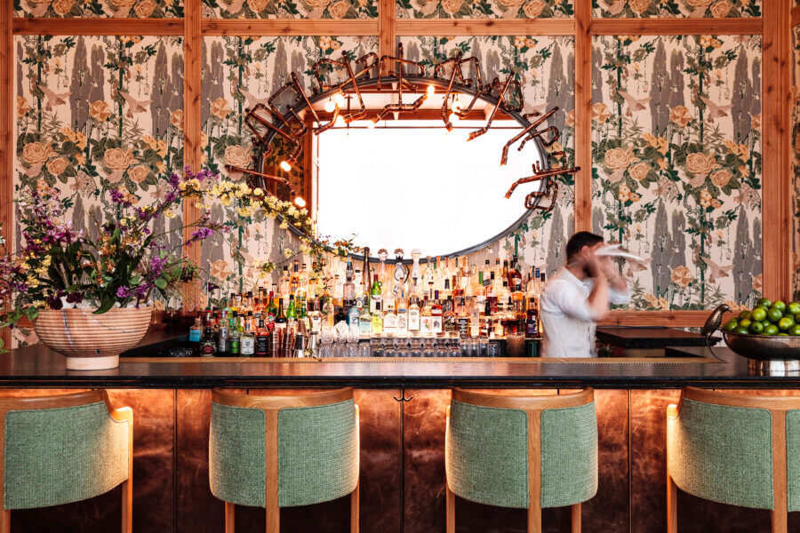 6 Striking Southern Hotels Designed by Top Talent - 4 A bartender is shaking a cocktail behind a bar with green chairs, floral wallpaper, a large decorative mirror, and shelves stocked with bottles.