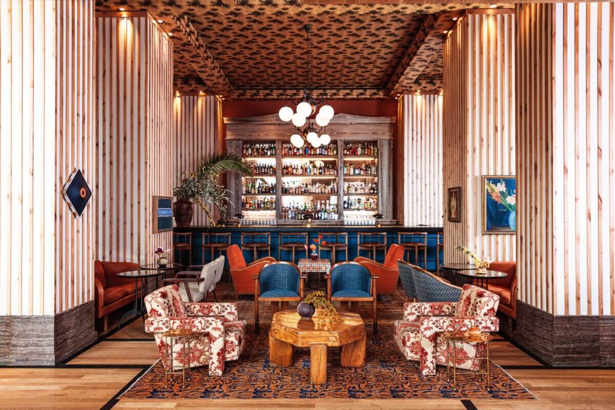 Eclectic lounge with floral armchairs, patterned rug, blue bar stools, wood-paneled walls, and a fully stocked bar in the background—showcasing Southern hotels' flair for inviting interior design. Geometric light fixture hangs from the ceiling.