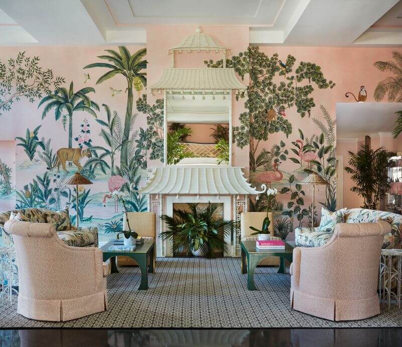 6 Striking Southern Hotels Designed by Top Talent - 5 A stylish pastel hotel space with four upholstered armchairs, green coffee tables, a fireplace with a pagoda-style mantle, and a tropical mural covering the pink walls.