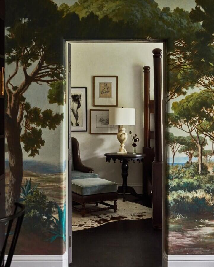 6 Striking Southern Hotels Designed by Top Talent - 14 A doorway with wall murals of trees opens to a room in one of the most beautiful hotels, featuring framed art, a lamp on a dark table, an upholstered chair, and a canopy bedpost visible.