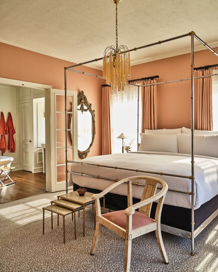6 Striking Southern Hotels Designed by Top Talent - 13 A bedroom with a gold canopy bed, white linens, a vintage chair, two small side tables, a large ornate mirror, and peach-colored walls and curtains.