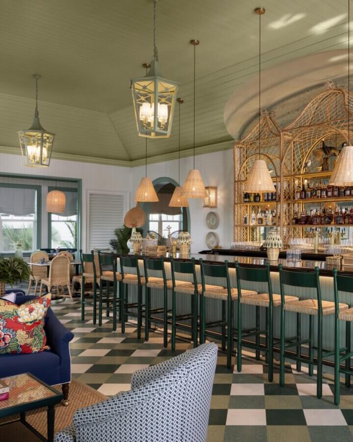 6 Striking Southern Hotels Designed by Top Talent - 7 Elegant bar area with high stools, wicker decor, checkered floor, and hanging lights. Shelves behind the bar display bottles. The room has a green ceiling and soft natural light—an ambiance found in the most beautiful hotels.