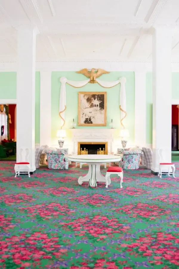 6 Striking Southern Hotels Designed by Top Talent - 12 A spacious room in one of the most beautiful hotels, featuring a floral carpet, white columns, pastel green walls, a fireplace, armchairs, and a round central table beneath a large framed painting.
