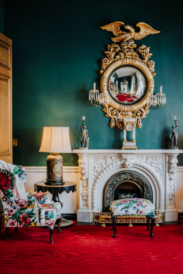 6 Striking Southern Hotels Designed by Top Talent - 10 Elegant room with a marble fireplace, ornate circular mirror, floral armchair and ottoman, a lamp, and deep green walls with red carpet—showcasing classic interior design often found in stylish hotels.