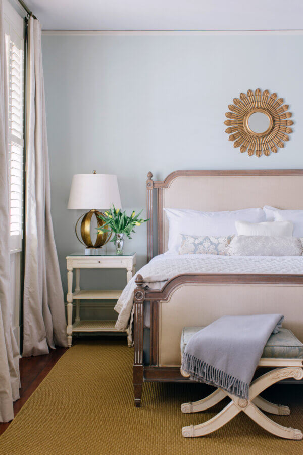 6 Striking Southern Hotels Designed by Top Talent - 17 A neatly made bed with neutral bedding sits next to a white nightstand, lamp, and vase of flowers; a sunburst mirror hangs on the light blue wall above.