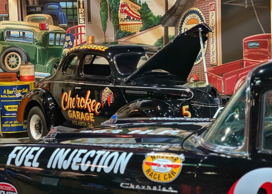 How to Eat, Shop & Explore Your Way Through Georgia - 14 A vintage black race car with "Cherokee Garage" branding is displayed indoors with its hood open, surrounded by other classic cars and automotive memorabilia—a must-see for fans searching for unique things to do in Georgia.