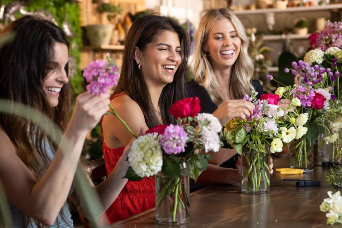 Three women sit at a table arranging colorful flower bouquets in glass vases, smiling and enjoying the activity—a perfect scene for girlfriend getaways in the South as they bond and create memories for 2026.