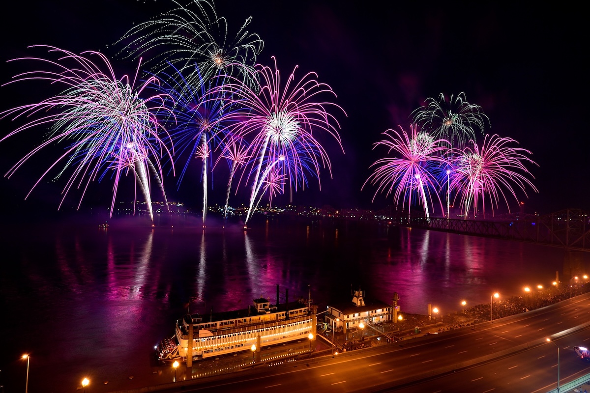 Fireworks in shades of pink, purple, and white light up the night sky above a river with a bridge, city lights, and a docked riverboat below during Louisville events in April.