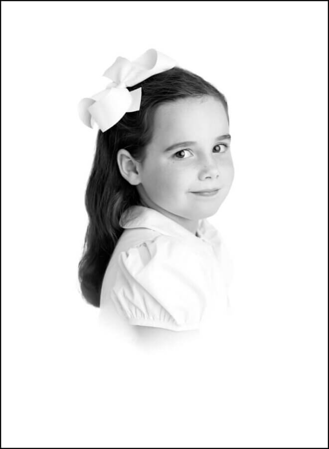 Heirloom Portraits Are Making a Comeback! - 3 Black and white portrait by Southern heirloom portrait photographers of a young girl with long dark hair, wearing a large white bow and a short-sleeved blouse, gazing slightly over her shoulder.