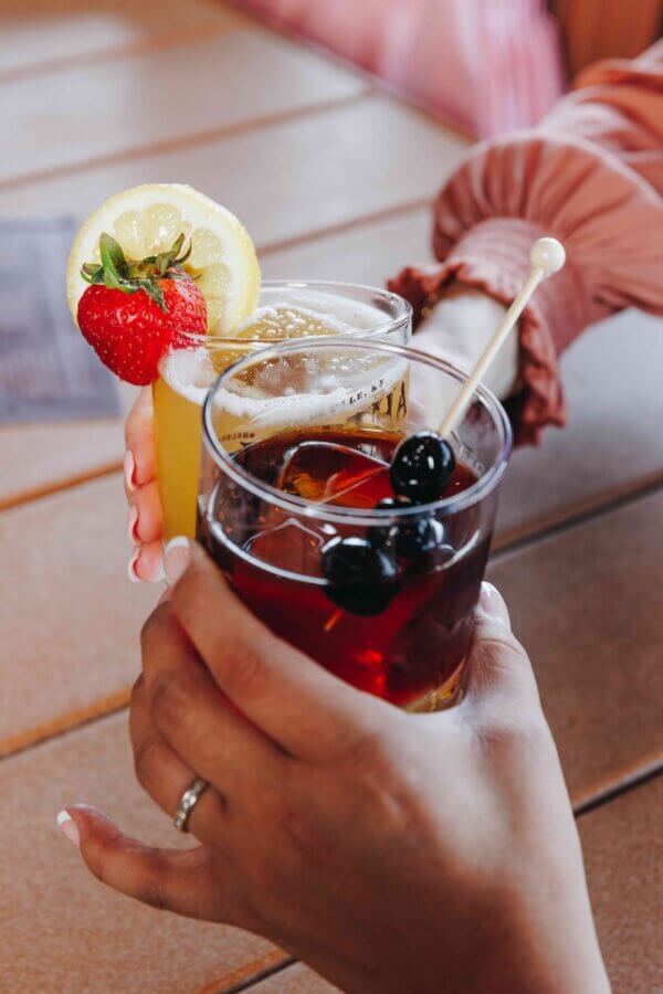 9 Hidden Gems to Visit in the South - 14 Two people clinking glasses, savoring hidden gems: one with a yellow drink garnished with lemon and strawberry, the other with a dark cocktail adorned with black cherries.