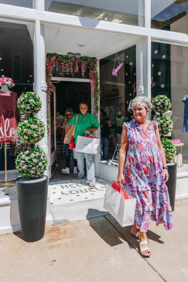 9 Hidden Gems to Visit in the South - 15 Three people exit a boutique store with shopping bags, exploring hidden gems; one woman in a floral dress walks ahead on the sidewalk while two others follow behind through the door.