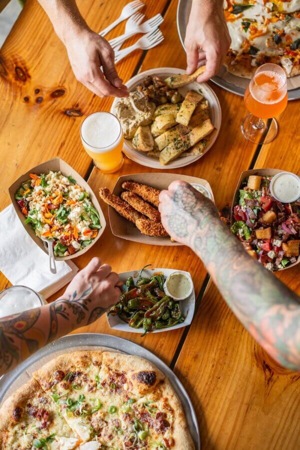 9 Hidden Gems to Visit in the South - 5 Hands reach for assorted food—including pizza, breadsticks, fried chicken, salad, and drinks—on a wooden table with plates, napkins, and utensils.